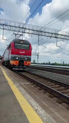 A passenger train moves along a track at a station. The train is red and gray with the logo of Russian Railways on the side. The train is moving from left to right in the frame, and the camera is positioned on the platform. The platform has a yellow line along the edge. The sky is cloudy, and there are overhead power lines for the train.