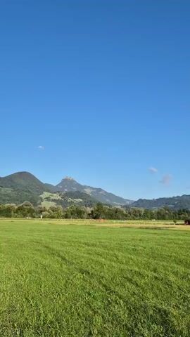 The video shows a vast, lush green meadow under a clear blue sky. In the distance, a range of mountains forms the backdrop, with some of the peaks showing rocky outcrops. There are a few buildings scattered on the landscape, including what appears to be a farmhouse with a dark roof and a shed-like structure. Trees are visible near the buildings and on the slopes of the mountains. The camera pans slowly from left to right, offering a panoramic view of the serene countryside. There are no people or animals visible in the frame. The lighting suggests it is daytime, likely morning or afternoon, with the sun casting soft shadows.