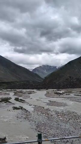 A video shows a wide river flowing through a valley. The riverbed is filled with stones and gravel, and the water is murky. On either side of the river are steep, barren mountains. The sky is overcast with thick, gray clouds. In the distance, snow-capped mountain peaks are visible. There are no people or any obvious signs of human activity in the video, aside from what appears to be a railing in the foreground.