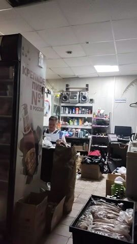 The video depicts the interior of a cluttered convenience store. A middle-aged person is standing behind a counter, appearing to be working. The store is filled with various items including boxes, bags, shelves stocked with products, and a stereo system. A large beverage cooler stands to the left, and a sign featuring beer is prominently displayed on it. The overall impression is one of disorder and a lack of organization.