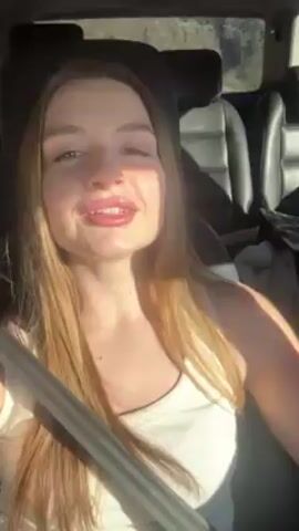 A young woman with long, light-brown hair is sitting in the driver's seat of a car. She wears a white tank top and a seatbelt. Her nails are painted red. She is singing or talking, gesturing with her hands. The car appears to be in motion. The background visible through the car windows is blurred but appears to be a wooded or mountainous area. The lighting is bright, suggesting it is daytime.