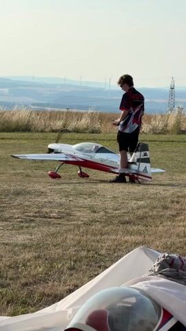 A young male wearing a red and black shirt and black shorts stands next to a remote-controlled airplane. The airplane is white with red accents and has its propeller spinning. The male is holding a remote control in his hands and appears to be operating the airplane. The scene is outdoors on a grassy field with a hazy landscape in the background. In the distance, there are wind turbines and a power line tower. The sky is clear and bright.