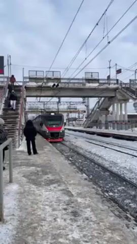 The video shows a train approaching a train station in winter. The train is gray and white with red and orange accents. It is marked "ЭП3Д-0017." The station platform is covered in snow. A few people are waiting on the platform, and others are walking up the stairs of a pedestrian bridge over the tracks. The sky is overcast.