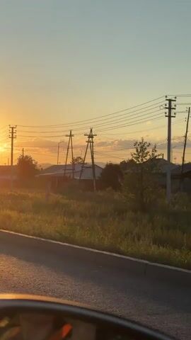 A video captures a view from inside a vehicle at sunset. The sun, a bright orb, is low on the horizon, casting a warm glow across the scene. In the foreground, a portion of the vehicle's dashboard is visible, and the asphalt road stretches ahead, reflecting the sunlight. To the left, a triangular road sign with a circular sign below it indicating a speed limit of 40 is positioned. Beyond the road, a grassy embankment is illuminated by the setting sun. In the middle ground, houses with pitched roofs and utility poles with wires are scattered across the landscape. The sky is a clear gradient of orange and blue, with a few wispy clouds. The overall impression is of a peaceful, rural setting during a golden hour.