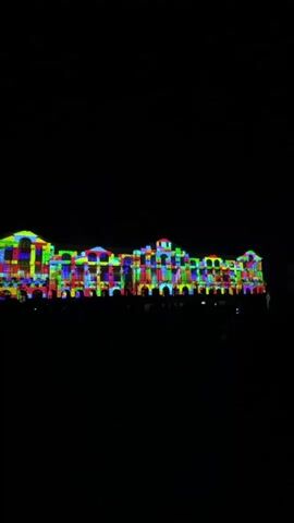 A wide shot captures a building at night, illuminated by a projection mapping display. The building facade is covered in a mosaic of brightly colored blocks, resembling large LEGO bricks. The colors include red, blue, green, yellow, purple, and orange, arranged in a grid-like pattern that alters the appearance of the windows and architectural features. In the foreground, a crowd of silhouetted people stands, facing the building, many holding up their phones to record the spectacle. The sky is completely dark, providing a stark contrast to the vibrant display on the building. The projection is dynamic, with colors and patterns shifting and moving across the facade, creating a lively and festive atmosphere. The scene appears to be an outdoor event or festival, possibly a public art installation.