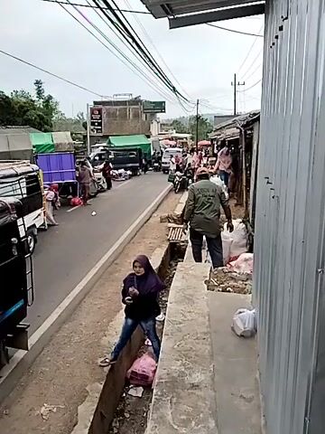 The video depicts an outdoor scene, likely a market or roadside with commercial activity. A two-lane road runs horizontally across the frame, with vehicles and people present on both sides. On the left side of the road, a white truck with a covered cargo area is parked, and several people, including a woman wearing a headscarf and jeans, are near it. Further down the road on the left, more vehicles, including a large green truck, and individuals are visible, with what appear to be market stalls or goods laid out. The road itself shows signs of wear and tear.

On the right side of the road, there's a ditch filled with debris and some pink plastic bags. A person wearing a dark jacket and a headscarf is leaning over the ditch, seemingly interacting with something within it. Behind this person, a narrower, unpaved walkway or area runs parallel to the road, separated by the ditch. Along this walkway, structures that appear to be stalls or shops are visible, constructed with corrugated metal and wood. Several people are present in this area, some interacting with goods that look like sacks of produce or other items. One individual in a green jacket is seen bending down and handling white sacks. Above the scene, a network of electrical wires is strung across the frame. The sky is overcast, suggesting a cloudy day. The overall impression is of a busy, informal commercial area with limited infrastructure.