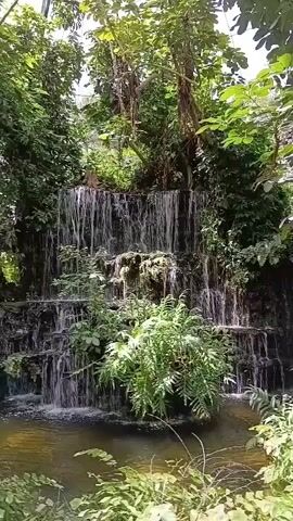 This is a vertical video of a man-made waterfall with multiple tiers. Lush green vegetation, including trees and ferns, surrounds the waterfall. The water flows down the tiers and collects in a pool at the bottom. The lighting suggests it is daytime. There are no people visible in the video.