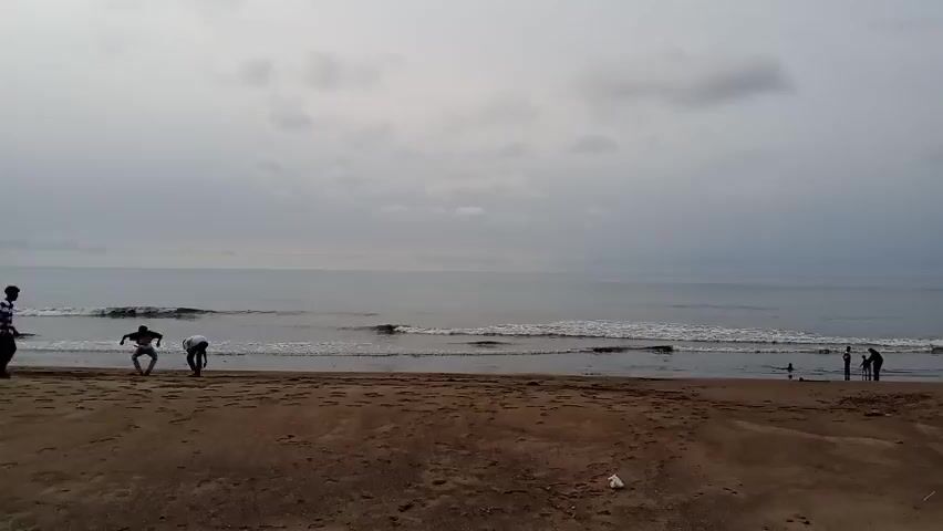 The video captures a wide shot of a beach with overcast skies. The ocean in the background exhibits gentle waves breaking on the shore. On the sandy beach, there are several individuals scattered across the frame. In the foreground and to the left, two people are visible, one appearing to be bending down as if picking something up. Further to the right, a group of three people, including what seems to be two adults and a child, are standing near the water's edge. Another individual is also present near them. The beach itself is sandy and appears to be mostly empty except for these few people. There is a piece of white litter visible on the sand in the lower center of the frame.