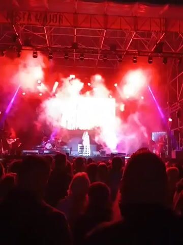 The video shows a female singer performing on stage during a concert. She is wearing a pink outfit and standing on a raised platform with lights. Behind her is a large screen displaying an image of trees at sunset. Red and pink stage lights illuminate the scene, and there is smoke or haze in the air. The audience is visible in the foreground, silhouetted against the stage. There are several musicians and instruments visible on the stage.