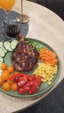 A top-down, panning shot showcases a dinner table setting. A green plate holds a grilled steak, sliced cucumbers, cherry tomatoes, red bell pepper strips, shredded carrots, and corn kernels. To the left are two glasses, one holding a golden liquid and the other a dark liquid. A decorative white ceramic rabbit and a gold candlestick with a white candle are placed on a dark gray woven placemat. The table has a light wood grain finish. The background shows a dark colored couch.