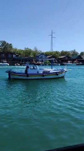 A small white and blue boat with a yellow stripe is moving through clear blue water. The boat has a canopy and appears to have at least one person on board. In the background, there are several buildings with dark roofs, surrounded by green trees. A tall transmission tower with wires is visible against a bright blue sky. The water is rippling, suggesting the boat is in motion.