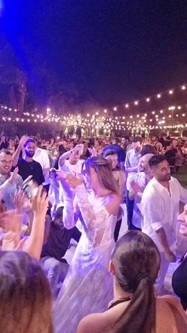 A group of people are dancing at night outdoors. A wedding dress is visible on a woman in the center of the frame. String lights are visible in the background. There is a camera set up to the right of the dancers.