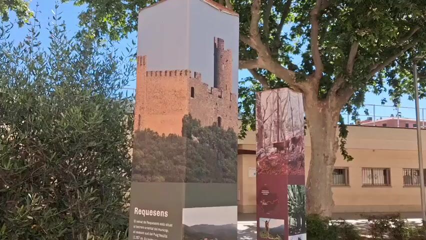 The video shows two tall, rectangular information kiosks standing on a paved area. The kiosk on the left has a photo of a castle on its upper section, and information about the town of Requesens on the lower part. The kiosk on the right has photos of hiking trails. A green bush is on the left side of the kiosk. A tree is on the right side of the kiosk. A building is visible in the background.