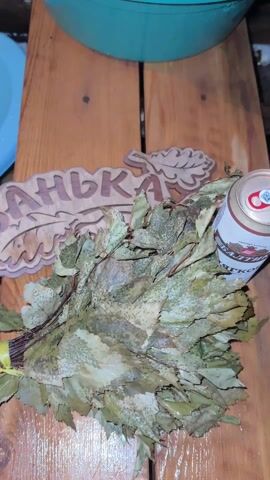 The video shows a wooden table with a leaf-shaped sign that says "Banja" (or "Banya") in Russian, a bundle of dried leafy twigs, and a can of drink. The camera zooms in to focus on the wooden sign, showcasing the dark brown lettering and leaf design. The scene suggests a traditional Russian sauna or bathhouse setting with related elements present. The video is captured from a close-up perspective with a slightly shaky camera movement.