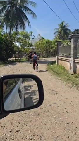 A group of children, each with a backpack, ride bicycles down a dusty, unpaved road lined with trees and simple structures. The camera is positioned inside a vehicle, showing a side mirror in the foreground, indicating that the vehicle is following the children. The sun is shining, creating a bright, outdoor scene.