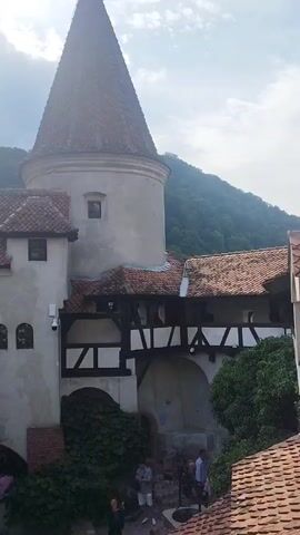 The video shows a courtyard of a castle, possibly Bran Castle in Romania, with a prominent conical tower. In the foreground, there is a dark wooden balcony with white railings, covered in ivy. People are visible on the balcony, some looking out. Below the balcony, there is an arched entranceway with a sign above it. Several people are walking in and out of the entranceway. The castle walls are white with red-tiled roofs. In the background, a lush green, forested hillside is visible under a cloudy sky. The lighting suggests it is daytime, possibly late afternoon due to the sun's angle and lens flare. The camera appears to be handheld, with slight movements and occasional wobbling.