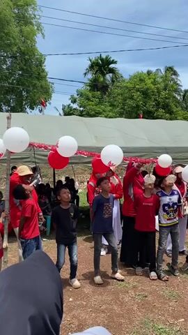 A group of children and adults are outdoors participating in a game. The children are standing in a line, facing forward, and looking upwards at a string of red and white balloons. Some of the children have small objects, possibly food items, attached to their faces, suggesting they are trying to eat them without using their hands. The adults, mostly dressed in red, are standing behind the children, some holding the balloons or supervising the game. The environment appears to be a somewhat informal outdoor gathering with a tent or canopy set up in the background. There are trees and vegetation visible, and the sky is bright and partly cloudy. The scene suggests a festive or celebratory event, possibly a competition or a cultural activity.