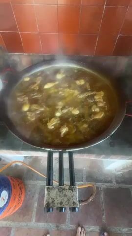 The video shows a wide, shallow pan full of stewing food being prepared over a three-pronged gas burner. The camera quickly shifts to follow a shirtless man wearing blue swim trunks and flip-flops as he walks across a brick-tiled patio. Another man in a white shirt is standing at a table with plates. A brown dog lies on the patio next to a white planter with pink flowers and a pink trash can. The video ends as the man walks toward a group of people seated at a table under a tree in the background.