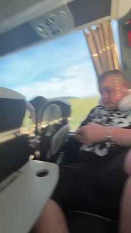 The video shows the interior of a bus, likely a tour bus, filled with passengers. Most passengers are seated, though a few are seen standing or moving about. Some passengers are drinking from cans.  The seats are black and silver, with fold-down trays. Overhead compartments run along the length of the bus. The windows show an exterior view of a sunny, possibly rural, landscape. The video pans and zooms, focusing on different passengers and the interior of the bus.