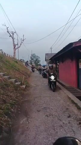 The video shows a group of people riding motorcycles on a narrow, unpaved road. The road is bordered by a grassy slope on the left and a building with a red wall on the right. Several motorcycles are visible, some with riders and some without. The sky is overcast, and the lighting suggests it is either early morning or late afternoon. The overall impression is that of a casual group ride through a rural or semi-rural area.