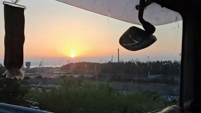 The video shows a view from inside a vehicle looking out a window at a sunset. The sun is low on the horizon, casting a warm glow across the sky. In the foreground, there is a rearview mirror hanging from the window, and a decorative item hanging from the top left of the window. Outside the window, there are trees and foliage. In the middle ground, there is a large parking lot filled with many cars, and beyond that, there are industrial structures and what appears to be a Ferris wheel in the distance. The overall impression is one of a scenic sunset over an industrial or commercial area. The camera appears to be moving, as evidenced by the motion blur in some parts of the video, particularly in the trees and foliage.