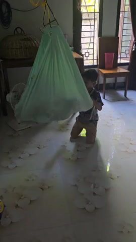 A young boy kneels on the floor beside a light green fabric baby swing, which hangs from a hook attached to a wooden structure. The boy is wearing a dark t-shirt with a design and brown pants. The swing contains a baby, though only a small portion of its body is visible. The room is lit by natural light coming through a window in the background. There are several white, petal-shaped objects scattered on the floor around the boy and the swing. A fan is visible on the left side of the frame, and a table with some items on it is in the background. The boy appears to be looking into the swing.