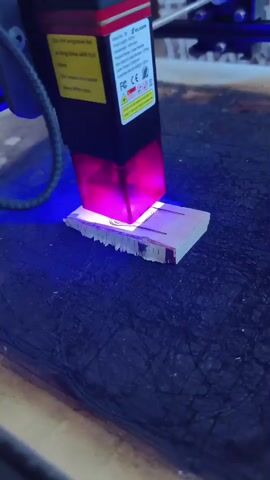 A close-up, low-angle shot shows a laser engraving tool positioned above a small piece of wood. The laser head, a black rectangular device with a red top and a yellow warning label, emits a bright pink and blue light onto the wood. The wood appears to be light-colored and roughly rectangular, with visible grain and some signs of being worked on, possibly being cut or etched. The laser is actively engraving a design onto the surface of the wood. The background is dark and textured, likely a workbench or a similar surface, with a hint of machinery or equipment visible in the upper right corner. The scene is illuminated by the bright laser light, creating a stark contrast with the darker surroundings.