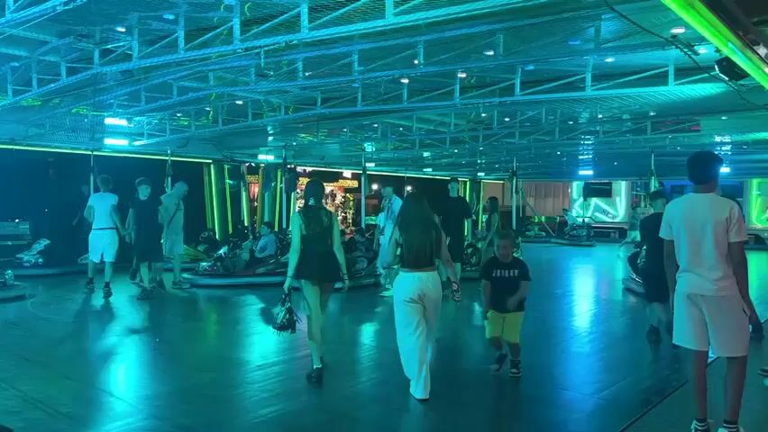 This is a video of a bumper car arena at what appears to be a fairground or amusement park. The scene is lit by bright blue and green neon lights, creating a vibrant and energetic atmosphere. Several people, mostly teenagers and young adults, are seen walking around the arena and preparing to ride the bumper cars. Some are already seated in the cars, looking excited and ready to go.

The arena has a polished, reflective floor that mirrors the colorful lights. The bumper cars themselves are brightly colored and have a classic design. In the background, there are signs and structures typical of an amusement park, including what looks like a ticket booth or concession stand. The overall mood seems festive and fun, with people dressed in casual summer clothing, suggesting it's likely a warm evening. The location context provided indicates this is in Riccione, Italy. There is no discernible weather information, but the indoor setting with artificial lighting makes it irrelevant. The only visible text is on a sign in the background that appears to read "PICCO" or something similar, possibly part of the name of the ride or attraction.