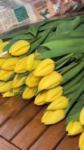 The video shows a bunch of yellow tulips lying on a wooden table. The tulips are densely packed, with their green leaves overlapping. A partially visible white paper bag with a floral design is in the background. The camera angle is slightly high, looking down at the tulips.