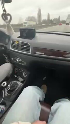 A passenger records a video from inside a moving car. The car is an Audi, with brown leather seats, black dashboard, and wood trim.  The driver is wearing gray sweatpants, a green jacket, and tan shoes.  The driver is holding a wad of US currency with their right hand. The view out the windshield shows other cars and trees on the side of a highway. The weather is cloudy and overcast. The car has an air freshener hanging from the rear view mirror. The person is listening to music in the car.