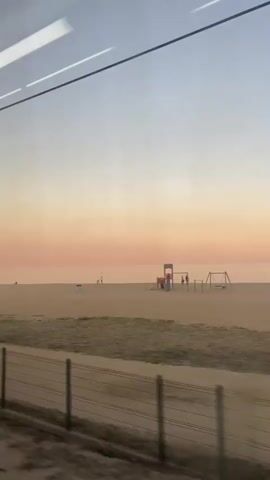 The camera is inside a moving train and looking through a window at a beach. In the distance, the sky is a gradient of light pink to pale blue. The sea is visible as a flat horizon line. The beach is wide and sandy, with some patches of grass along the fence line that separates it from the train tracks. There are people walking on the beach, including a couple, two women, and a group of people standing together. There is also a small playground structure on the beach. The train is moving quickly, causing the scene to pass by rapidly. The interior of the train is visible in the reflection on the window, including overhead lights and wires. The fence is made of metal posts and horizontal bars.