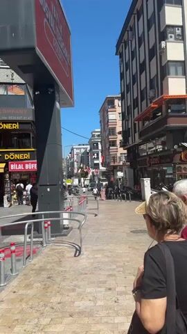 The video opens with a view of a street scene on a sunny day. In the foreground, two people, an older woman with dark hair wearing a black t-shirt and a long, floral skirt, and an older man with grey hair wearing a red t-shirt and grey shorts, are walking away from the camera. They are holding hands. The woman is carrying a black bag over her shoulder. The man is carrying a dark strap over his shoulder. They are walking on a paved sidewalk with metal railings. To their left is a large, black, rectangular sign with a red rectangle at the top. Further to the left, there are shops and buildings. One shop has a yellow sign that reads "BAYBURT DONER". A red trolley car is visible on the left side of the frame, stopped at a red area on the street. The trolley has "100" written on it and "bir binicilik" which translates to "one ride". Several people are visible in the background, walking on the street and sidewalks. The buildings are multi-story and appear to be shops and businesses. The sky is clear and blue. The camera pans slightly to the right, following the couple. More buildings and shops are visible. The trolley car continues to be present on the left. The people in the background are less distinct. The overall impression is of a busy urban street.