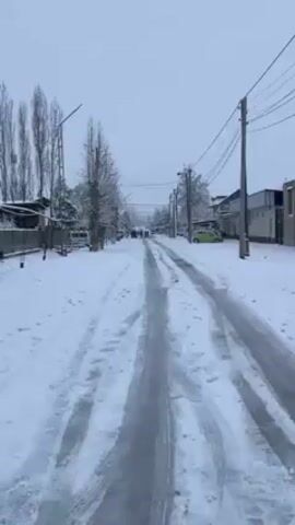The video shows a street covered in snow. Tire tracks run down the middle of the road. On either side of the road are buildings, trees, and power poles. In the distance, a group of people is walking toward the camera. Further down the road, two vehicles are parked. The sky is overcast.