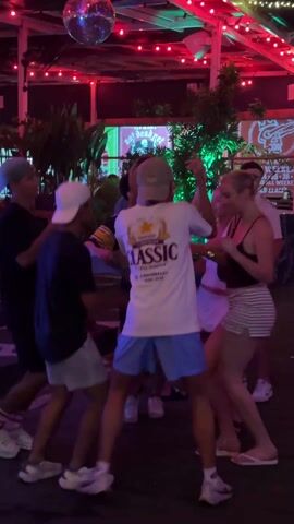 The video shows a group of six young adults, including three men and three women, dancing in a circle at night. A disco ball hangs above them, and red and blue lights illuminate the scene. The men are wearing casual clothing, including shorts, T-shirts, and baseball caps. One of the men is wearing a T-shirt with the word "Classic" on it. The women are also wearing casual clothing, including skirts, tops, and shorts. They appear to be having a good time as they dance and interact with each other in a lively outdoor setting.