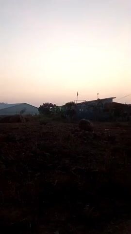 The video shows an outdoor scene at dusk. The foreground is a patch of dark, tilled soil with several large rocks scattered across it. In the middle ground, there are a few trees and buildings silhouetted against the sky. The buildings appear to be residential, with some having visible lights. A flag is visible on one of the buildings. The sky is a gradient of pale yellow and white, indicating the setting sun or twilight. A small object, possibly an airplane or bird, is visible in the sky at 00:02.