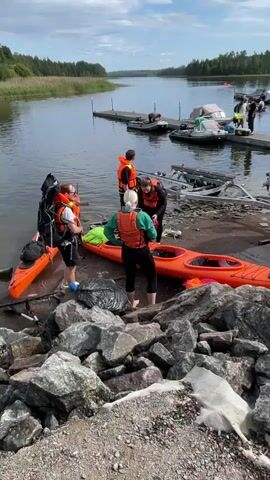 Four people are gathered near the water's edge, preparing to launch kayaks. Two orange kayaks are visible, one with a person in an orange life vest standing next to it, holding a large black backpack. Another person in a green shirt and orange life vest is kneeling beside a second orange kayak, appearing to adjust something. A third person in an orange life vest is also bending over the kayak. A fourth person, also in an orange life vest, stands slightly behind them. The kayaks are on a rocky shore, with a dock extending into the water behind them. Several other boats, including an inflatable dinghy, are moored at the dock. The environment is a natural setting with trees and vegetation along the far bank of the water body. The sky is cloudy.
