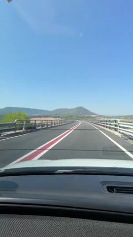 This is a first-person view from inside a car traveling on a highway bridge. The scene is set during the daytime, under a clear blue sky with no visible clouds, suggesting fair weather. The road is paved and has distinct lane markings, including a double white line and a prominent red stripe down the center.

The car is moving forward over a bridge with railings on either side. Beyond the bridge, a landscape of rolling hills covered in vegetation stretches into the distance. A single, conical-shaped mountain is visible as a prominent landmark against the horizon.

Several other vehicles are present on the highway. A white car is seen in the distance, and a red car is also visible. These vehicles indicate ongoing traffic on this road.

The overall mood is one of travel and a peaceful journey through a scenic, rural environment. The bright sunlight and clear skies contribute to a sense of openness and freedom. There is no visible text in the image. The location context is Manresa, Spain.