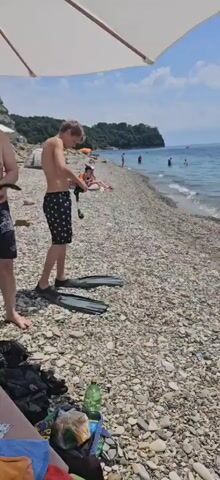 A teenage boy stands on a pebble beach wearing black swim trunks with white polka dots and swim fins. He is holding snorkeling gear in his hands. To his left, another person's legs are visible, wearing black shorts with white designs. In the background, the ocean is visible with several people swimming. The beach is covered in smooth, grey pebbles. A white umbrella is positioned overhead. The sky is blue with some clouds, and a green, tree-covered hill is visible in the distance.