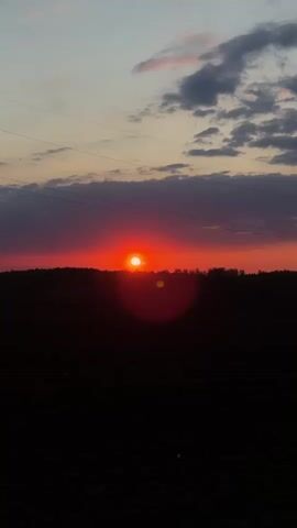 The video shows a bright red sun setting over a dark landscape, with a few puffy clouds in the upper right sky. There are power lines crossing the upper part of the frame. The landscape in the foreground is completely black, obscuring any detail. The sun has a noticeable lens flare.
