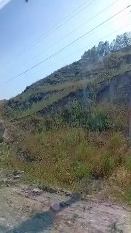 The video shows a view from a vehicle, looking uphill towards a rocky, grassy slope. Power lines are visible against a clear blue sky. The vegetation on the slope appears dry and sparse in places, with patches of green grass and some scattered bushes. The ground in the foreground is a dirt road with loose rocks. There are no people or animals visible in the frame. The sun is shining, casting shadows and creating glare on the lens.