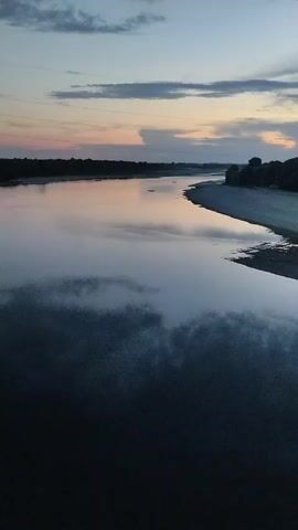 The video shows a slow pan across a river at dusk. The sky is a mix of blues and oranges, with some clouds. The water is calm and reflects the sky. The banks of the river are lined with trees. The overall impression is one of peace and tranquility.