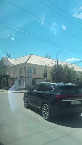 The video shows a street scene from inside a vehicle. A black SUV is driving away from the camera on a city street. In the background, there is a light-colored building with a light brown roof, trees, and traffic lights. A pedestrian crossing sign is visible. A digital display shows the temperature is 32 degrees Celsius. The sky is clear and blue. The video is shaky, indicating it was likely recorded from a moving vehicle.