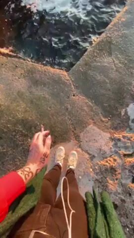 The video starts from a first-person perspective looking down at a person standing on a small, weathered concrete platform. The person is wearing tan sneakers, dark pants, and a red sweater, with a jacket over the sweater. Their right hand, adorned with henna-like designs, holds a cigarette. The camera slowly tilts up to reveal the ocean. Waves gently lap against the rocks and the concrete platform. In the distance, a vibrant sunset paints the sky with orange and golden hues. The water reflects the sunlight.  The camera continues to focus on the sea and the sunset, offering a panoramic view of the scenery.