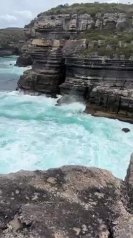 The video shows a coastal scene with rough seas crashing against layered rock formations. The water is a vibrant turquoise color with white foam from the breaking waves. The rock cliffs are dark gray and black, with green vegetation on top. The sky is overcast, and the camera is positioned on a rocky outcrop, offering a view down to the water.