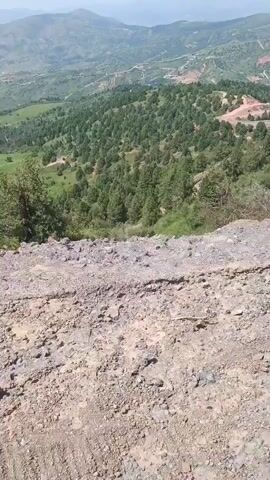 The video is a first-person perspective of someone looking down at a vast, mountainous landscape. The foreground is dominated by a rocky, dusty surface, likely a dirt road or embankment. Beyond this, the terrain slopes downwards, revealing a valley densely covered with evergreen trees, resembling a forest. The trees are primarily of medium height and a dark green color. Further in the distance, rolling hills and mountains stretch towards the horizon. The sky is a pale blue with some haze. The overall impression is of a remote, natural environment, possibly a rural or undeveloped area. There are no people, vehicles, or specific structures visible. The camera appears to be stationary, with the view focused on the expansive scenery.