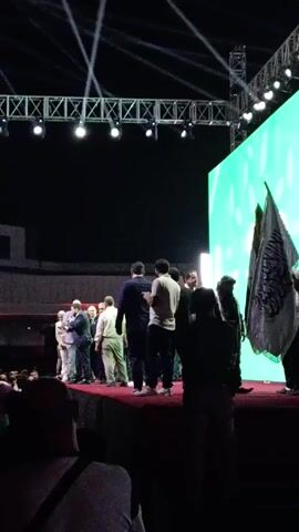 The video depicts a group of individuals on a stage, likely at an event or ceremony. The stage is covered with a red carpet, and there's a large screen in the background displaying a green abstract pattern. A lighting truss is visible overhead. The group on stage consists mostly of men in suits, with one woman in a light-colored pantsuit. Some individuals are standing, while others appear to be in conversation. A flag is visible on the right side of the stage. The camera captures the scene from the perspective of someone in the audience, with the heads and shoulders of other audience members visible in the foreground. The environment appears to be a large indoor space, possibly an auditorium or hall.