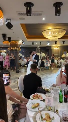 The video shows a wedding reception. A bride and groom are dancing in the center of the room, surrounded by guests seated at tables. The room is decorated with chandeliers and floral arrangements. Some guests are recording the dance with their phones. Smoke is visible on the dance floor, possibly from a special effect. The lighting suggests it is evening.