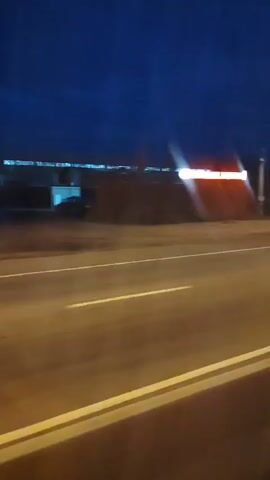 The video shows a blurry nighttime view from a moving vehicle. The scene is dominated by a dark blue sky and a road with white lane markings. On the right side of the road, there are streetlights with bright, streaking lights. Buildings are visible in the background, some with illuminated windows and signs. The primary action involves cars passing by, their headlights and taillights creating streaks of light due to the motion blur and the low light conditions. The overall impression is of a highway or main road at night, with traffic flow.