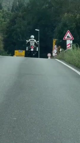 A motorcycle is being filmed from behind as it drives up a paved road surrounded by trees. A black SUV and a silver sedan are driving towards the motorcycle. Several road signs are visible, including a speed limit sign indicating 40, and a yellow directional sign. The sky is overcast.