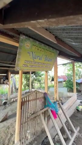 This media file shows an outdoor scene, likely in Pante Beurene, Indonesia, on a clear day with palm trees visible in the background. The central focus is a simple wooden structure with a corrugated metal roof that appears to be in disrepair, possibly a small mosque or prayer area.

Several people are present. Two men stand near the right side of the structure, engaged in conversation. One man, wearing a pink shirt and dark pants, gestures outwards, while another man in a white shirt and light brown pants looks in the same direction. Another individual, partially obscured, is seen in the background near a similar structure.

Inside the wooden structure, a sign is prominently displayed, reading: "MAJLIS TAKLIM 'Nurul Islam' PANTE BEUREUNE KUTA SIMPANG KEC. MEURAH DUA KAB. PIDIE JAYA". This translates to "Nurul Islam Taklim Council, Pante Beureune, Kuta Simpang, Meurah Dua District, Pidie Jaya Regency." The sign suggests the location is a place of religious gathering or education.

The ground around the structure is muddy and uneven, with scattered debris, indicating a rural or possibly a developing area. A small, fenced-off area within the structure, possibly a grave site, is covered with a colorful cloth. A wooden ladder leans against the enclosure. The overall impression is of a functional but humble community space, possibly undergoing construction or renovation, set against a tropical backdrop. The time of day appears to be late afternoon, judging by the lighting.